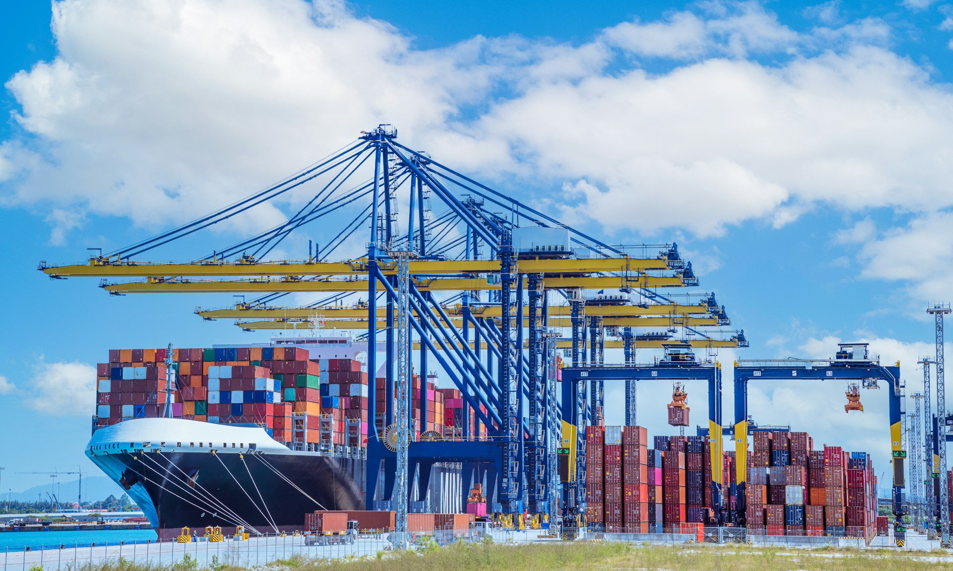 Image d'un port à conteneurs sous un ciel bleu avec des nuages ​​blancs. À gauche, un grand cargo est amarré, chargé de conteneurs colorés empilés. À droite, des grues jaunes et bleues sont en opération, soulevant et déplaçant des conteneurs. Le cadre véhicule une atmosphère d'activité et de logistique intenses, avec les infrastructures du port clairement visibles.